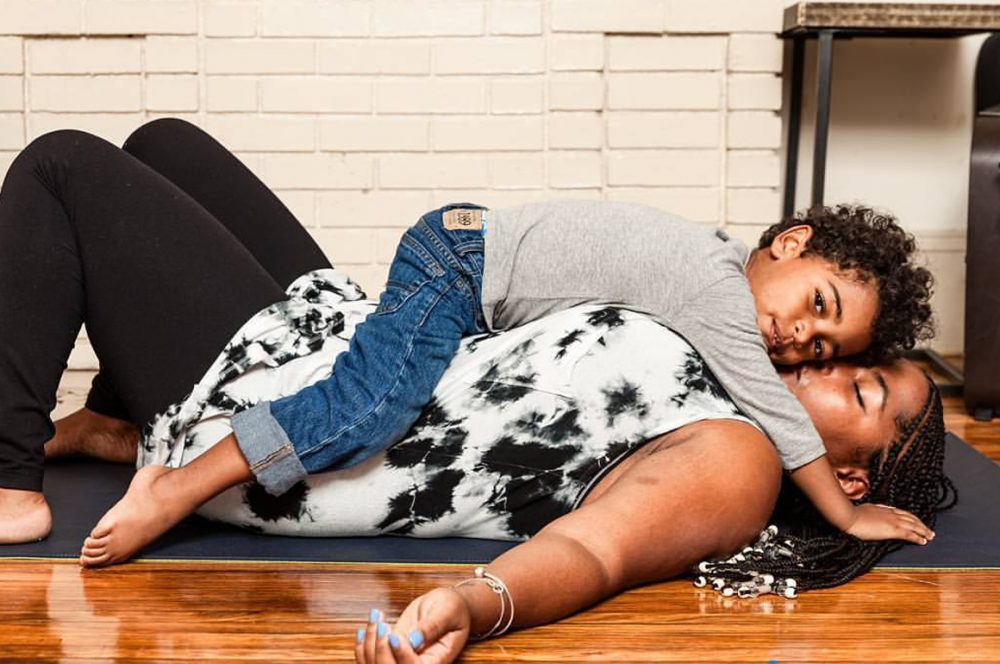Kelley, a Black woman, in Constructive Rest pose on a yoga mat on the floor on top of polished floorboards with her child lying over her torso. She is kissing him on the cheek.
