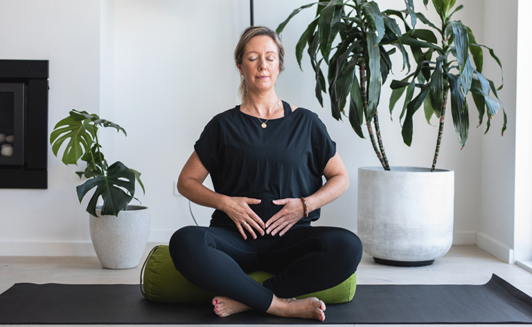 Tanya Neate, a white woman with blonde hair wearing a black top and black leggings sitting on a green bolster, legs crossed with hands on womb space. There are pot plants in the background.