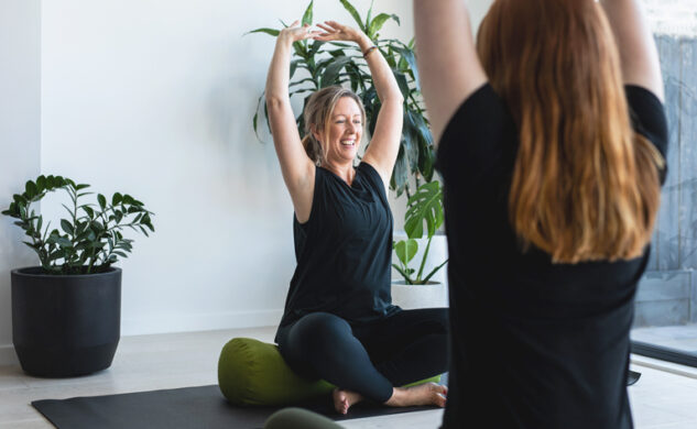 Tanya Neate, a blonde white woman, practicing yoga with hands clasped reaching overhead with a class participant with long auburn hair in the foreground