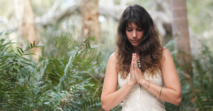 Rosie Matheson, a white woman with long brown wavy hair, with eyes closed and head tilted down, hands together at heart centre, with trees and greenery in the background