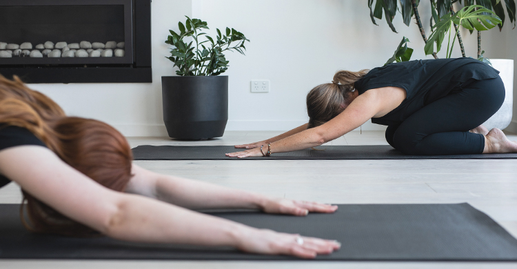 Two women in child's pose on yoga mats with plants in the background