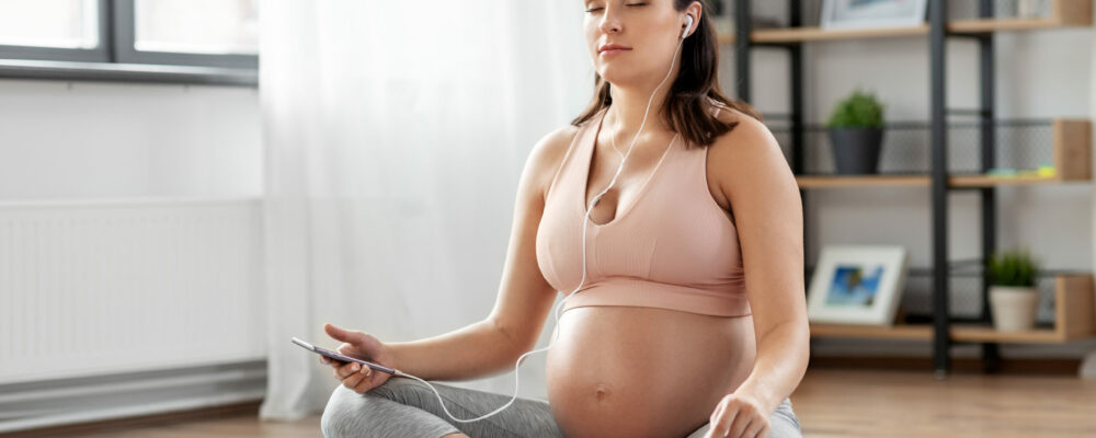sport, yoga and people concept - happy pregnant woman with earphones and smartphone listening to music and meditating at home